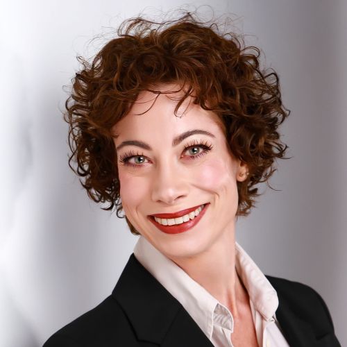 Porträt einer lächelnden Frau mit lockigem, braunem Haar, in einem schwarzen Blazer und weißem Hemd.