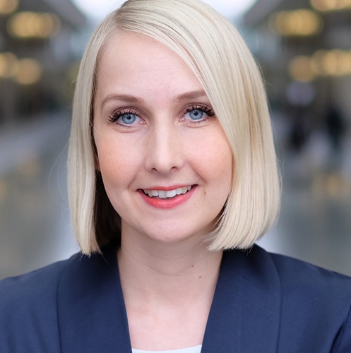 Porträt einer blonden Frau mit kurzem Haar, die in einem blauen Blazer lächelt, vor einem unscharfen Hintergrund.