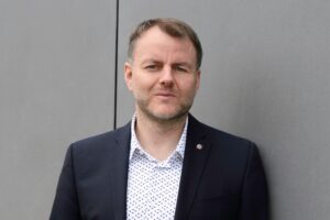 Mann in Anzug mit Hemd, steht an einer grauen Wand und schaut direkt in die Kamera.