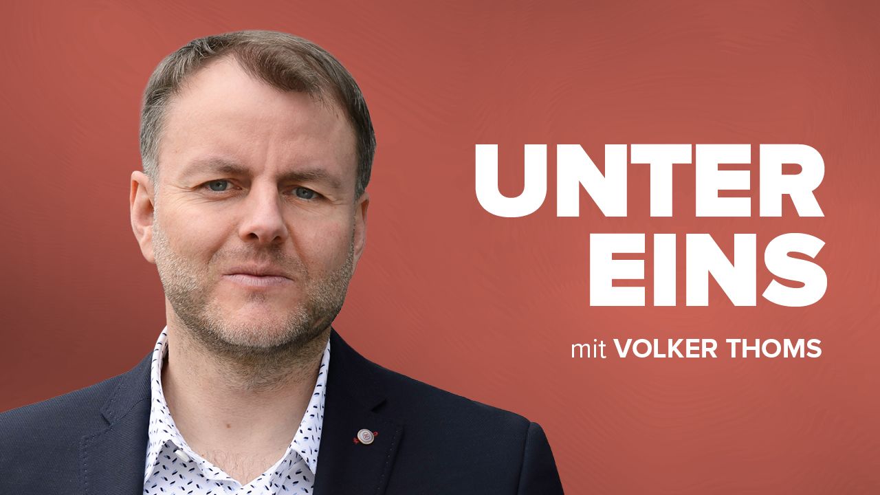 Mann mit kurzem Haar und Anzug steht vor rotem Hintergrund, Titel „UNTER EINS“ und „mit VOLKER THOMS“ in großer S...