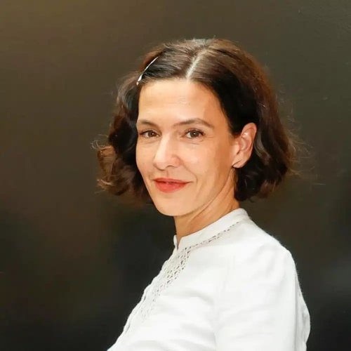 Frau mit schulterlangen, lockigen Haaren und einem weißen Oberteil, lächelt vor einem dunklen Hintergrund.