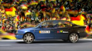 Blaues Auto mit Werbung für mobile.de und eBay Motors vor einer jubelnden Menschenmenge mit Deutschlandflaggen.
