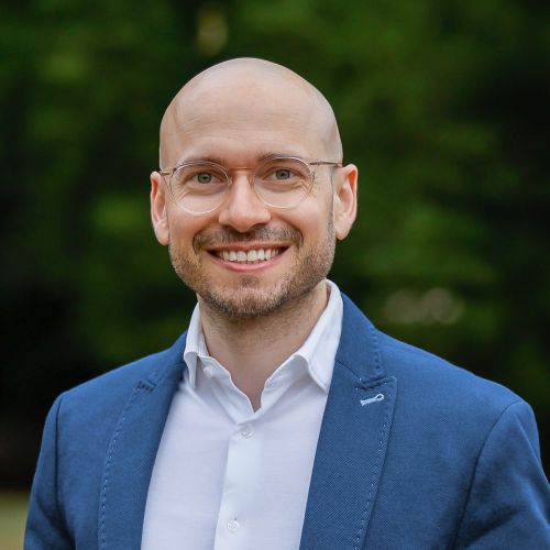 Porträt eines lächelnden Mannes mit Glatze, Brille und blauer Jacke, vor unscharfem grünen Hintergrund.