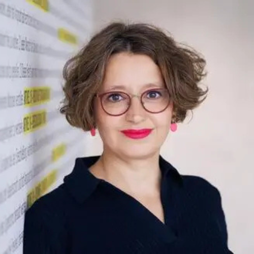Professional woman with short curly hair and glasses, wearing a dark blouse, standing beside a wall with highlighted notes in the background.