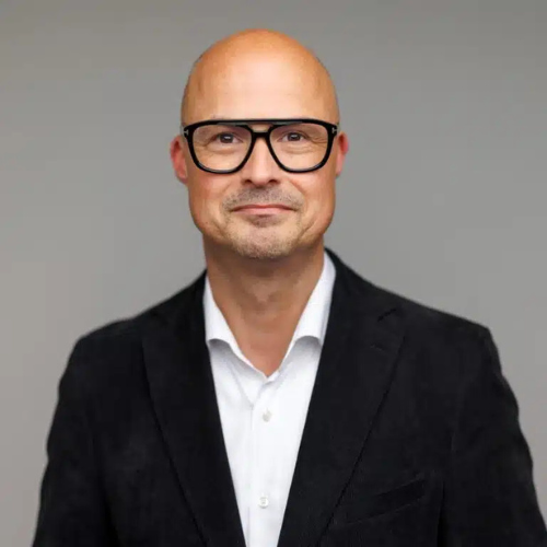 Portrait of a bald man wearing black-rimmed glasses, a white shirt, and a black blazer, standing against a neutral gray background.