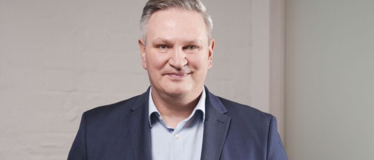 Mann mit grauen Haaren in Anzug und Hemd, lächelt vor einer hellen Wand.