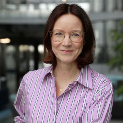 Frau mit Brille und gestreiftem Hemd lächelt in einem modernen Büro.