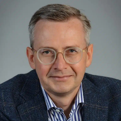 Porträt eines Mannes mit Brille, grauem Haar und einem blauen Blazer, lächelnd vor neutralem Hintergrund.