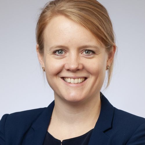 Porträt einer lächelnden Frau mit blonden Haaren, die einen blauen Blazer trägt, vor einem neutralen Hintergrund.
