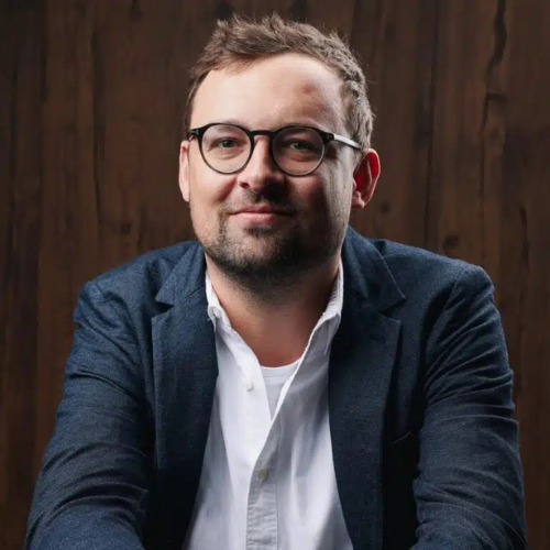 Mann mit Brille und Anzug, lächelt in die Kamera vor einer Holzpaneel-Wand.