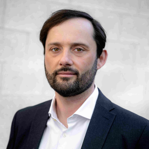 Professional headshot of a man with dark hair and beard wearing a navy suit and white shirt, looking at the camera.