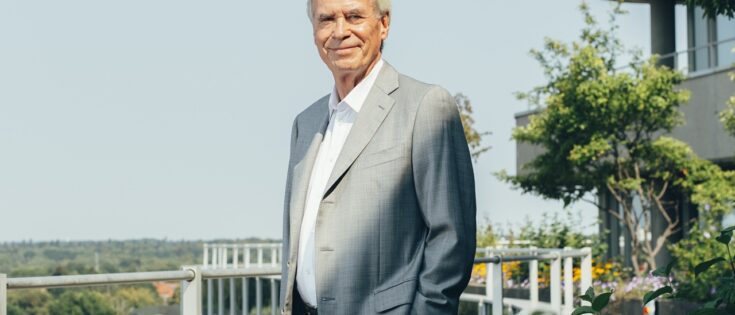 Portrait eines älteren Mannes in einem grauen Anzug, stehend auf einer Terrasse mit Pflanzen und Blick auf die Landschaft.