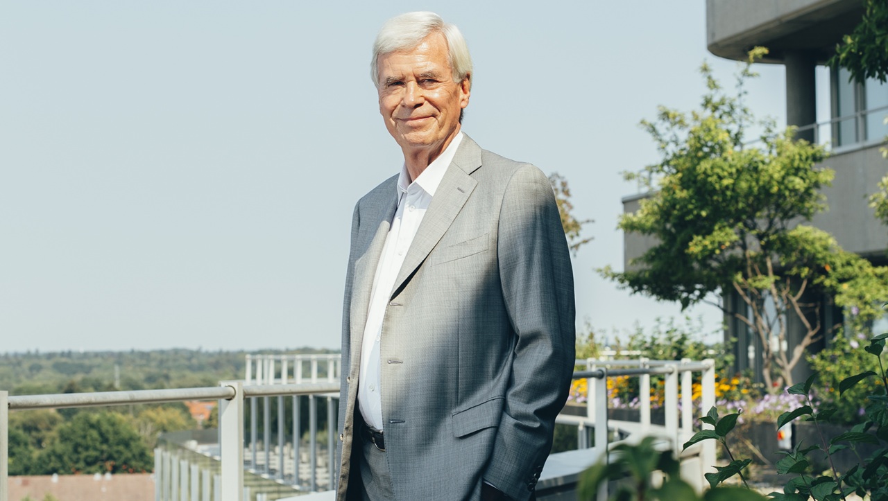 Portrait eines älteren Mannes in einem grauen Anzug, stehend auf einer Terrasse mit Pflanzen und Blick auf die Landschaft.