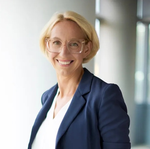 Frau mit blonden, kurzen Haaren und Brille, trägt einen blauen Blazer, lächelt vor einem hellen Hintergrund.