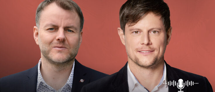 Volker Thoms und Mischa Heuer in Anzügen vor einem roten Hintergrund, mit einem Mikrofon-Symbol rechts.