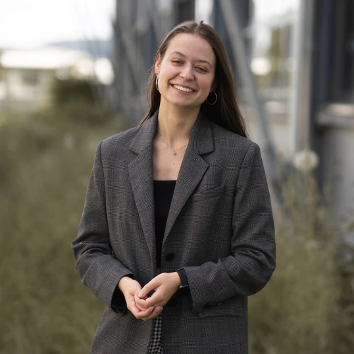Junge Frau mit langen Haaren in einem grauen Blazer lächelt vor einem modernen Hintergrund mit Pflanzen.