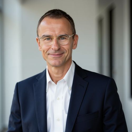 Mann mit Brille und Anzug lächelt vor einem neutralen Hintergrund.