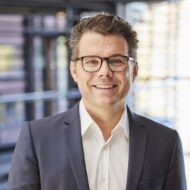 Mann mit Brille und Anzug lächelt vor modernem Hintergrund.