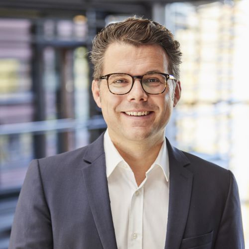 Mann mit Brille und Anzug lächelt vor modernem Hintergrund.