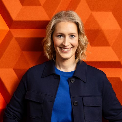 Anna-Maria Schuck lächelt vor einer orangefarbenen, geometrischen Wand und trägt eine dunkelblaue Jacke und ein blaues S...