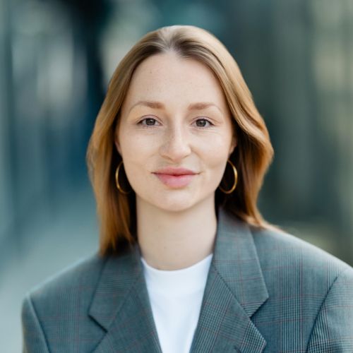 Porträt von Nina Weise mit blonden Haaren, in einem grauen Blazer, vor unscharfem Hintergrund.