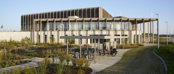 Moderne Gebäude mit Glasfassade, umgeben von einem Garten und Fahrradstellplätzen, unter blauem Himmel.