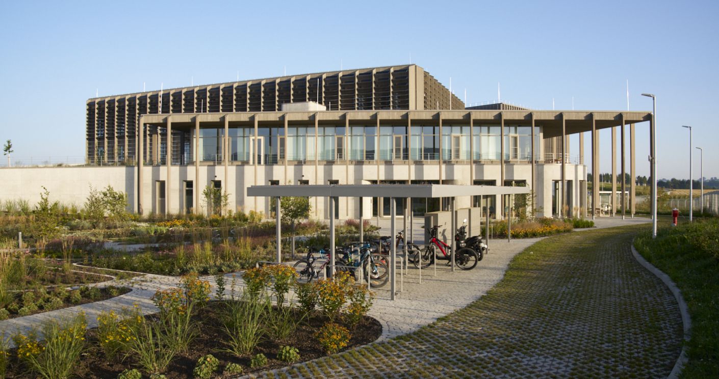 Moderne Gebäude mit Glasfassade, umgeben von einem Garten und Fahrradstellplätzen, unter blauem Himmel.