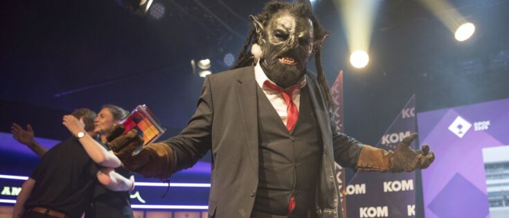 Ein Ork in Anzug mit Maske und Handschuhen steht auf einer Bühne, umgeben von bunten Lichtern und jubelnden Menschen.