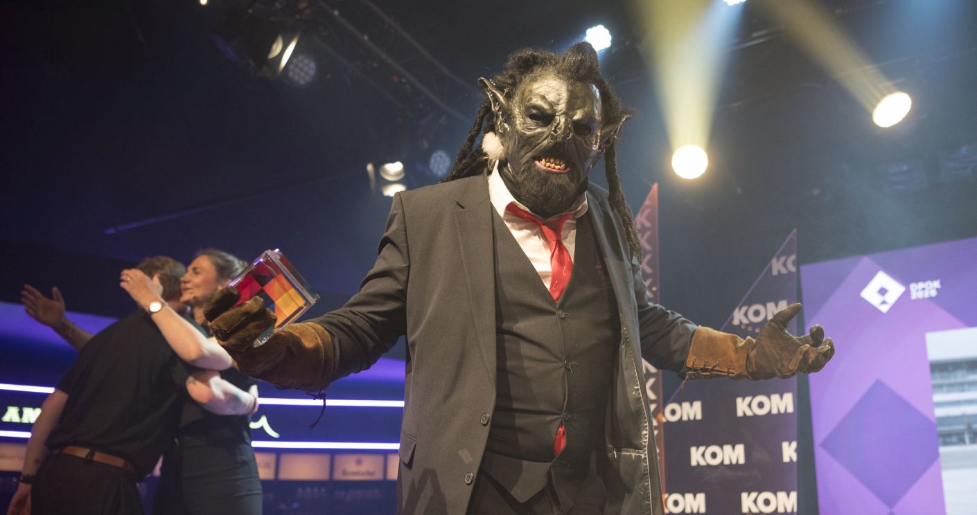 Ein Ork in Anzug mit Maske und Handschuhen steht auf einer Bühne, umgeben von bunten Lichtern und jubelnden Menschen.