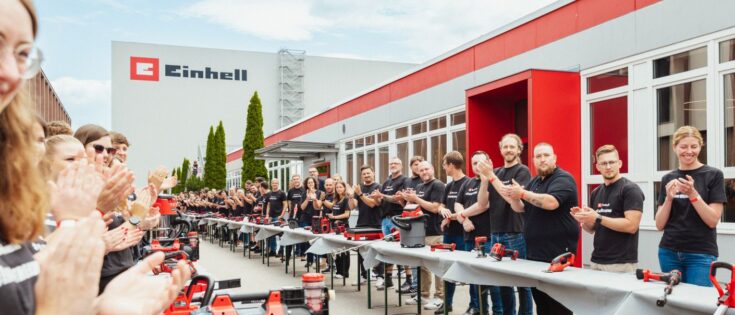 Gruppenszene mit Menschen in schwarzen Einhell-T-Shirts, die applaudieren, umgeben von Werkzeugen auf Tischen vor einem Einhell-Gebäude.