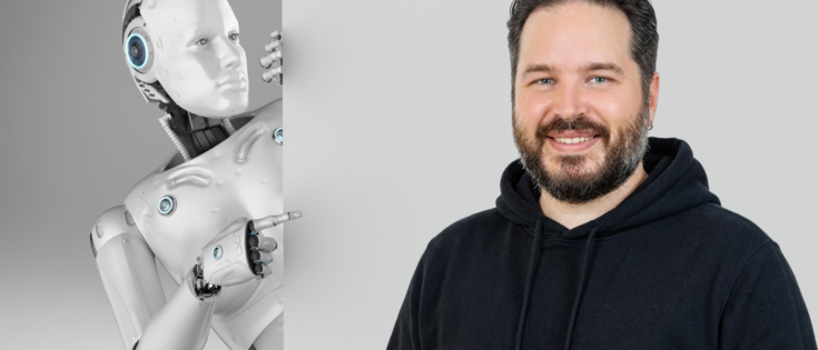 Ein humanoider Roboter zeigt auf einen lächelnden Mann in einem schwarzen Hoodie, vor einem grauen Hintergrund.