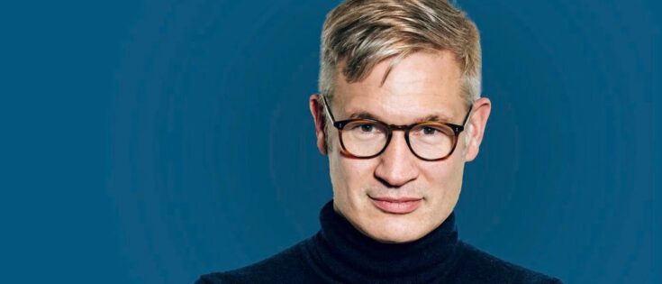 Mann mit Brille und kurzem, blondem Haar in einem schwarzen Rollkragenpullover vor blauem Hintergrund.