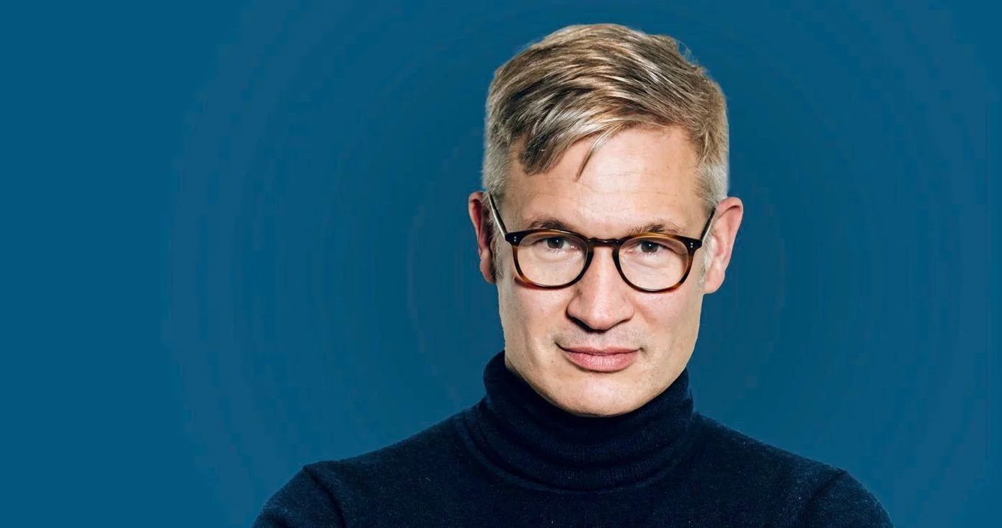 Mann mit Brille und kurzem, blondem Haar in einem schwarzen Rollkragenpullover vor blauem Hintergrund.