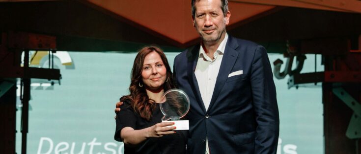 Zwei Personen stehen auf einer Bühne, eine hält einen Preis in der Hand, im Hintergrund eine grüne Leinwand.