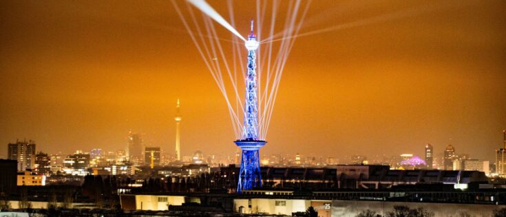 Funkturm in Berlin, beleuchtet mit blauen Lichtern und Strahlen, vor einer städtischen Skyline bei Nacht.