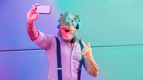 Ein tättowierter Mann mit T-Rex-Maske macht ein Selfie vor einem bunten Hintergrund.