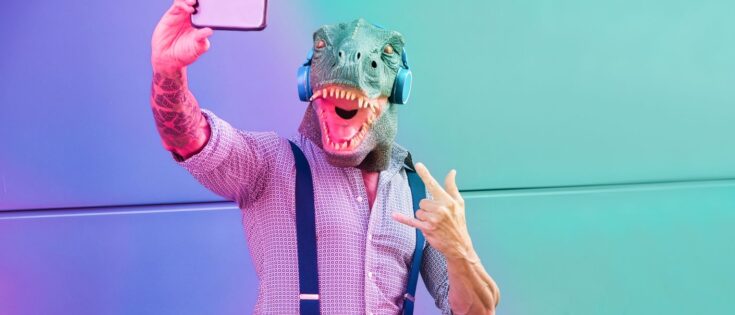 Ein tättowierter Mann mit T-Rex-Maske macht ein Selfie vor einem bunten Hintergrund.