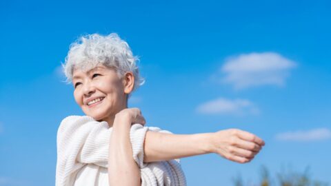 Seniorin mit weißen Locken dehnt sich lächelnd unter blauem Himmel mit weißen Wolken.