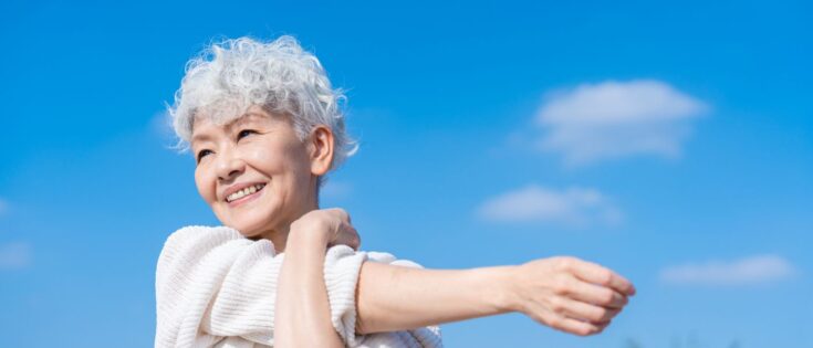 Seniorin mit weißen Locken dehnt sich lächelnd unter blauem Himmel mit weißen Wolken.