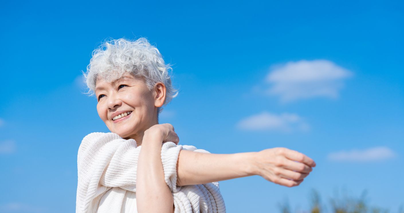 Seniorin mit weißen Locken dehnt sich lächelnd unter blauem Himmel mit weißen Wolken.