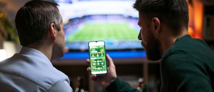 Zwei junge Männer schauen ein Fußballspiel im Fernsehen und nutzen gemeinsam eine mobile Sportwetten-App.