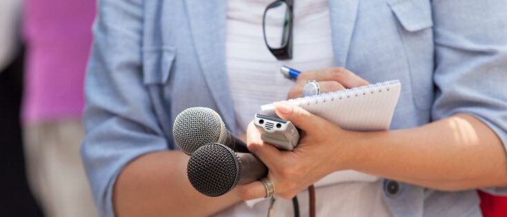 Journalistin hält Mikrofone und Notizblock während einer Pressekonferenz.