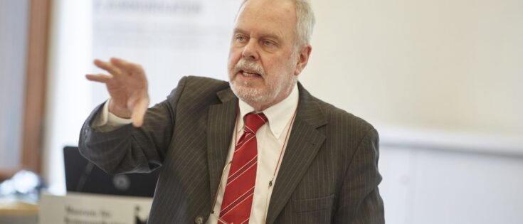 Älterer Herr mit weißem Bart und Anzug mit roter Krawatte hält gestikulierend einen Vortrag.