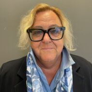 Porträt von Uta Hellweg mit blonden, lockigen Haaren, Brille und einem blauen Schal, vor grauem Hintergrund.