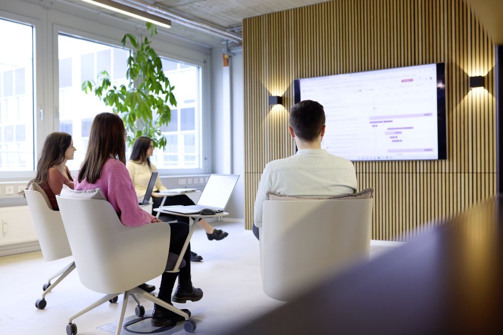 Vier Personen in einem modernen Besprechungsraum, die auf einen Bildschirm mit Präsentation schauen. Pflanzen im Hintergr...