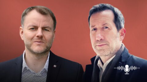 Zwei Männer stehen vor einem roten Hintergrund, einer in Anzug, der andere in einem Mantel, beide schauen in die Kamera.