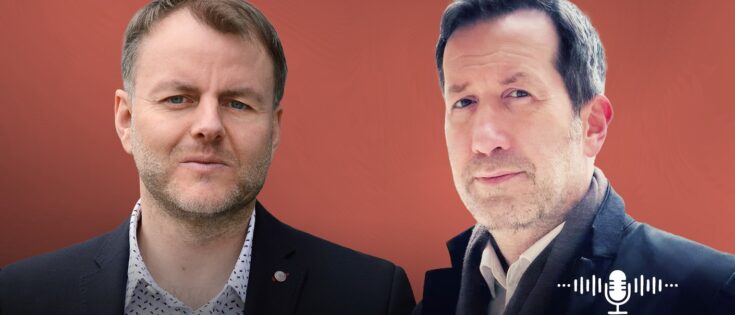 Zwei Männer stehen vor einem roten Hintergrund, einer in Anzug, der andere in einem Mantel, beide schauen in die Kamera.