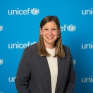 Portrait von Charlotte van Randenborgh vor einem blauen Hintergrund mit dem UNICEF-Logo, lächelnd in einem grauen Blazer.