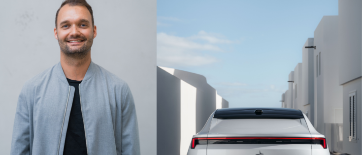 Mann in grauer Jacke lächelt, neben einem weißen Auto vor modernen Gebäuden unter blauem Himmel.