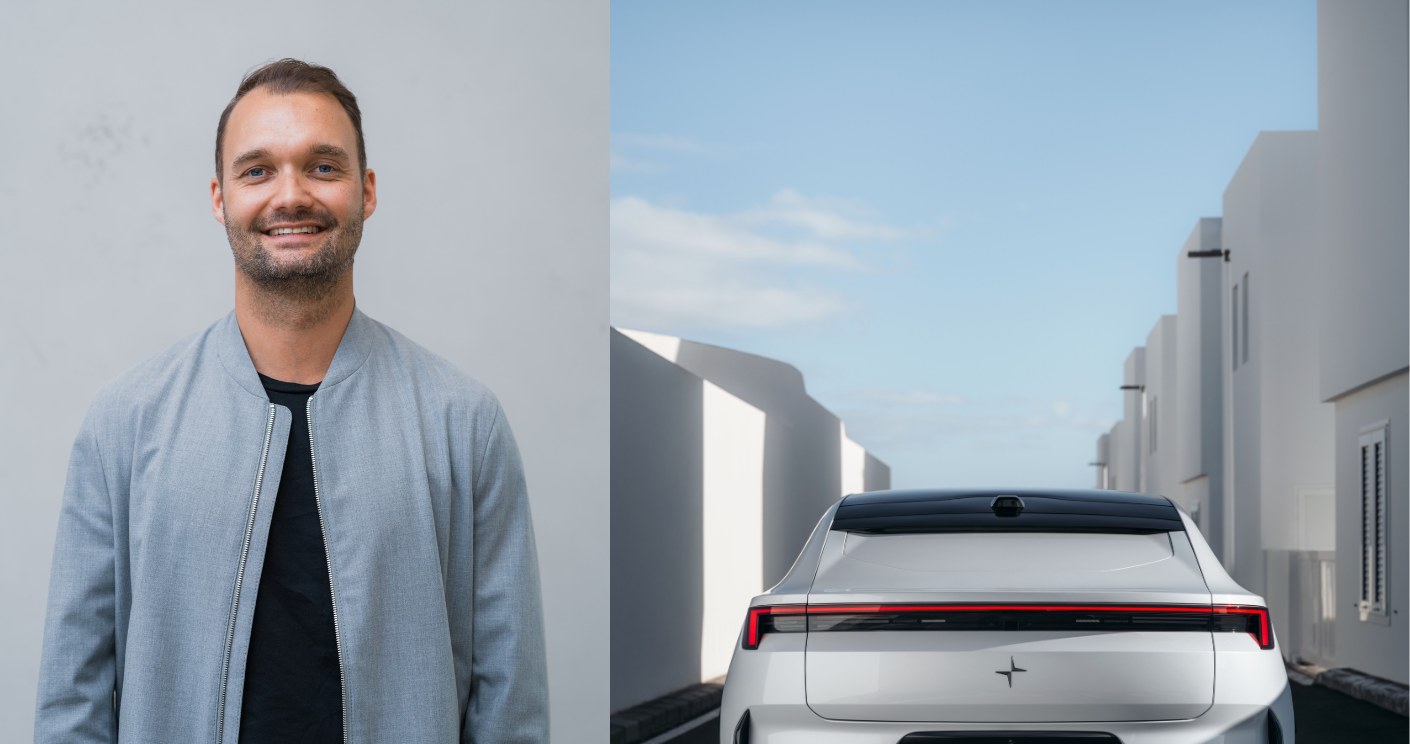 Mann in grauer Jacke lächelt, neben einem weißen Auto vor modernen Gebäuden unter blauem Himmel.